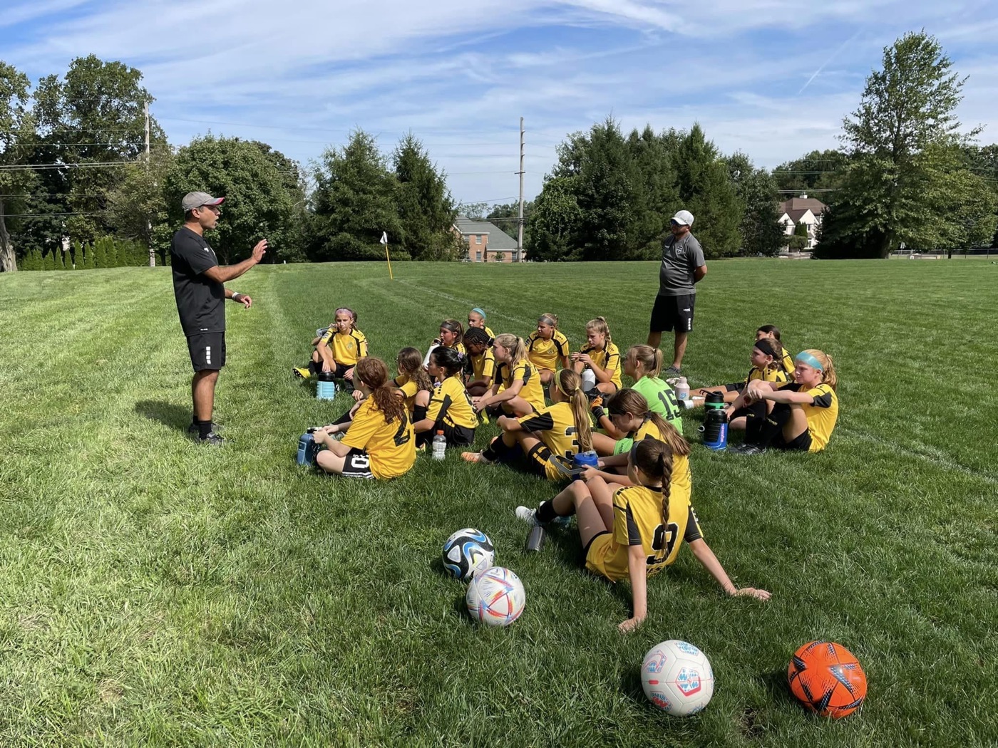 A Deptford girls team in gold kits sitting on the grass listening to their coach